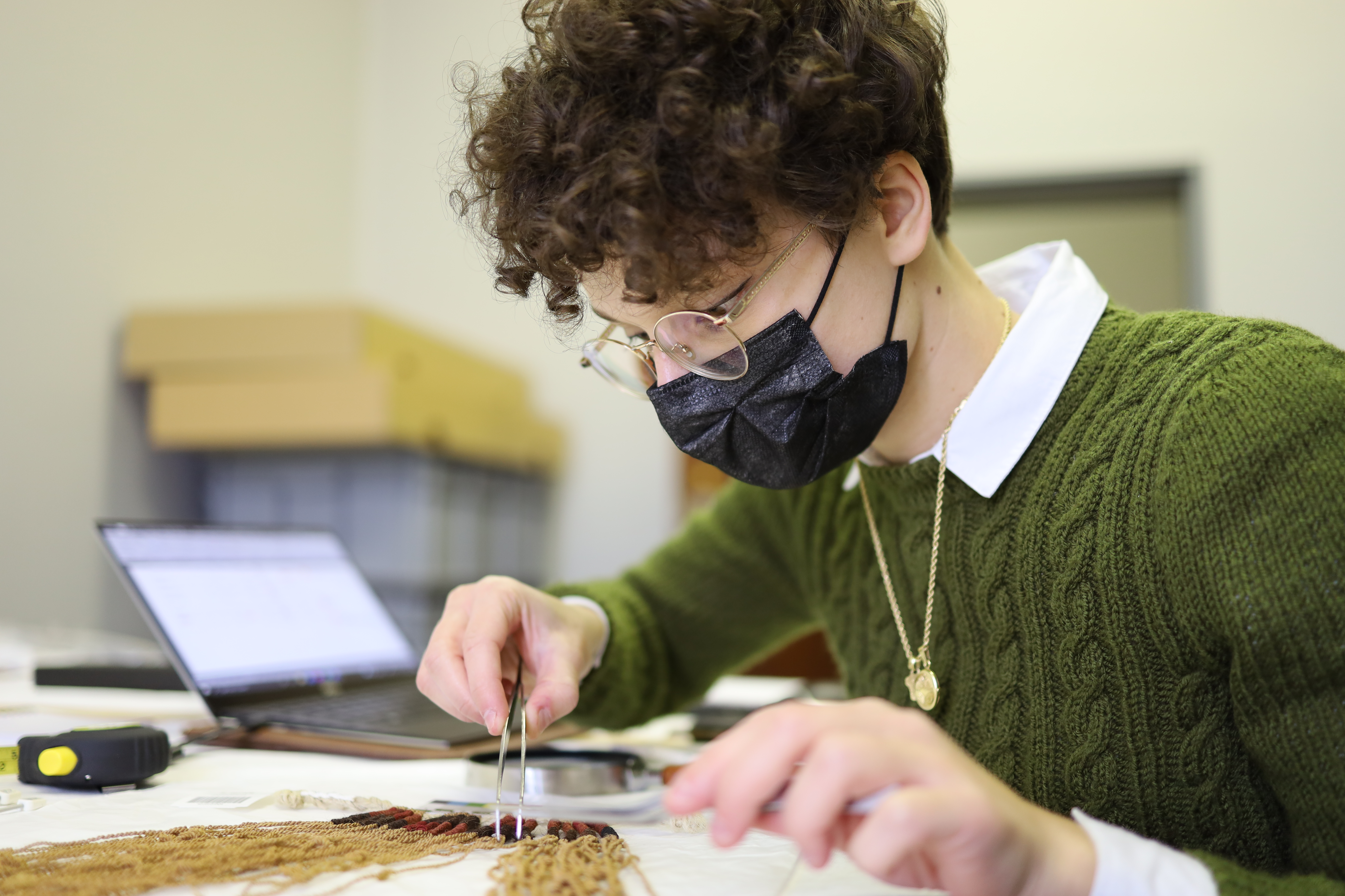 Lucrezia Milillo studying a khipu. The khipu is laying on a flat surface and she is approaching it with tweezers and magnifying glass. She is wearing a mask